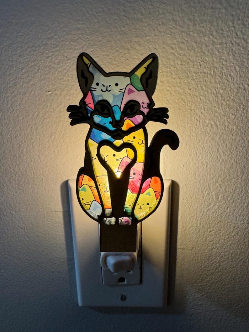May include: A colorful cat night light plugged into an outlet. The cat is made of black plastic and has a multi-colored design with a heart in the center. The light is on and illuminating the cat.