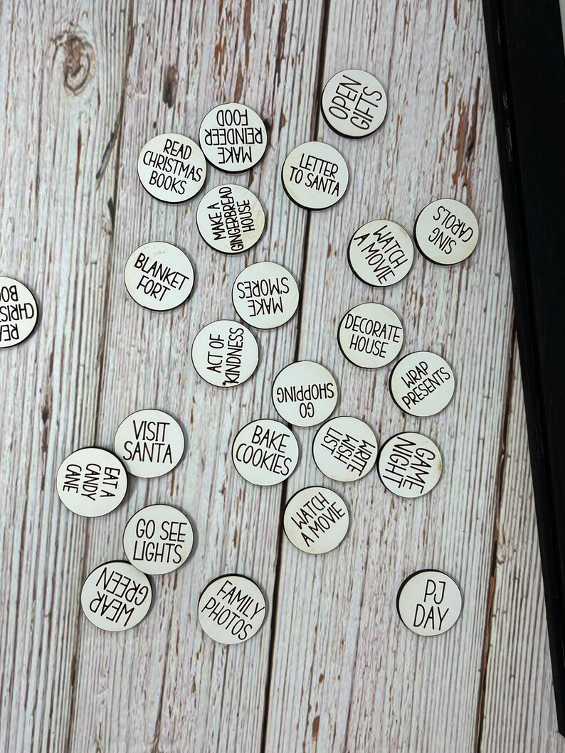 May include: A set of 20 white wooden circles with black text, each circle featuring a different Christmas activity. The activities include "Make Food", "Make S'mores", "Watch a Movie", "Decorate House", "Visit Santa", "Go See Lights", "Family Photos", "Bake Cookies", "Game Night", "PJ Day", "Read Christmas Books", "Open Gifts", "Letter to Santa", "Blanket Fort", "Act of Kindness", "Wrap Presents", "Sing Carols", "Eat Candy Cane", "Wear Green", "Write Wish List".