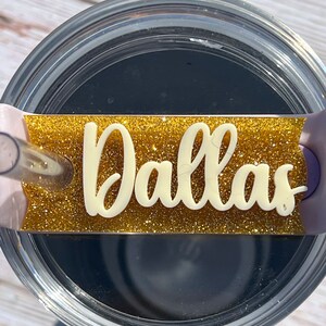 Puede incluir: Un vaso transparente con una tapa de purpurina dorada y una etiqueta de vinilo blanco que dice "Dallas".