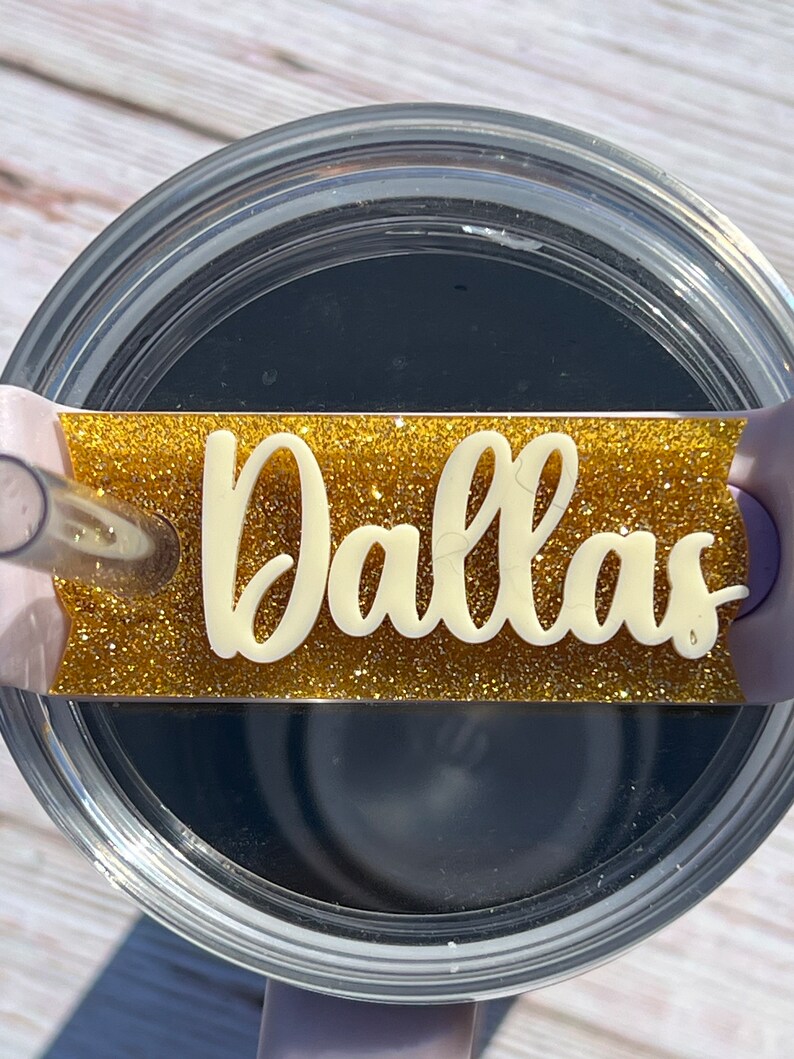 Puede incluir: Un vaso transparente con una tapa de purpurina dorada y una etiqueta de vinilo blanco que dice "Dallas".