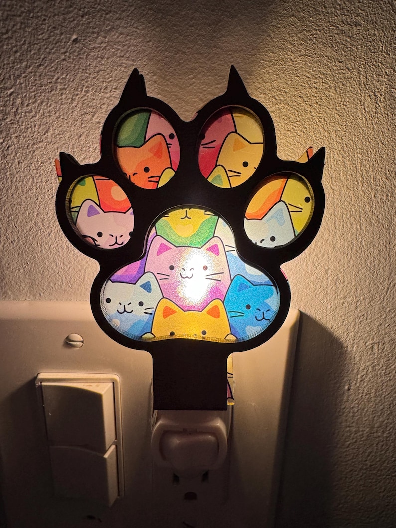 May include: A black paw-shaped night light with colorful cat faces inside each pad. The light is plugged into a wall outlet and emits a soft glow, creating a whimsical and decorative accent for a room.
