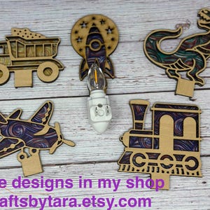 May include: Wooden night light cutouts in the shapes of a dump truck, rocket, dinosaur, airplane, and train. The cutouts have a layered design with colorful swirls. A night light bulb is in the center. Text at the bottom reads "More designs in my shop Craftsbytara.etsy.com."