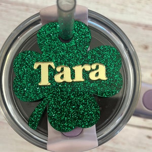 Puede incluir: Un tr&eacute;bol de purpurina verde con el nombre "Tara" en letras doradas. El tr&eacute;bol est&aacute; unido a la tapa de un vaso de acero inoxidable.