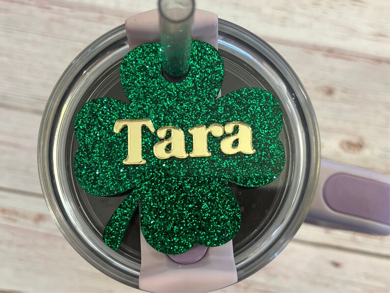 Puede incluir: Un tr&eacute;bol de purpurina verde con el nombre "Tara" en letras doradas. El tr&eacute;bol est&aacute; unido a la tapa de un vaso de acero inoxidable.