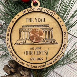 May include: Wooden Christmas ornament with a penny at the center, featuring the text "IN GOD WE TRUST" and "THE YEAR." The ornament includes the text "WE LOST OUR CENTS 1793-2025" and "DUE TO HIGH COST OF PRODUCTION, THE LAST PENNY WAS MINTED NOVEMBER 12, 2025."