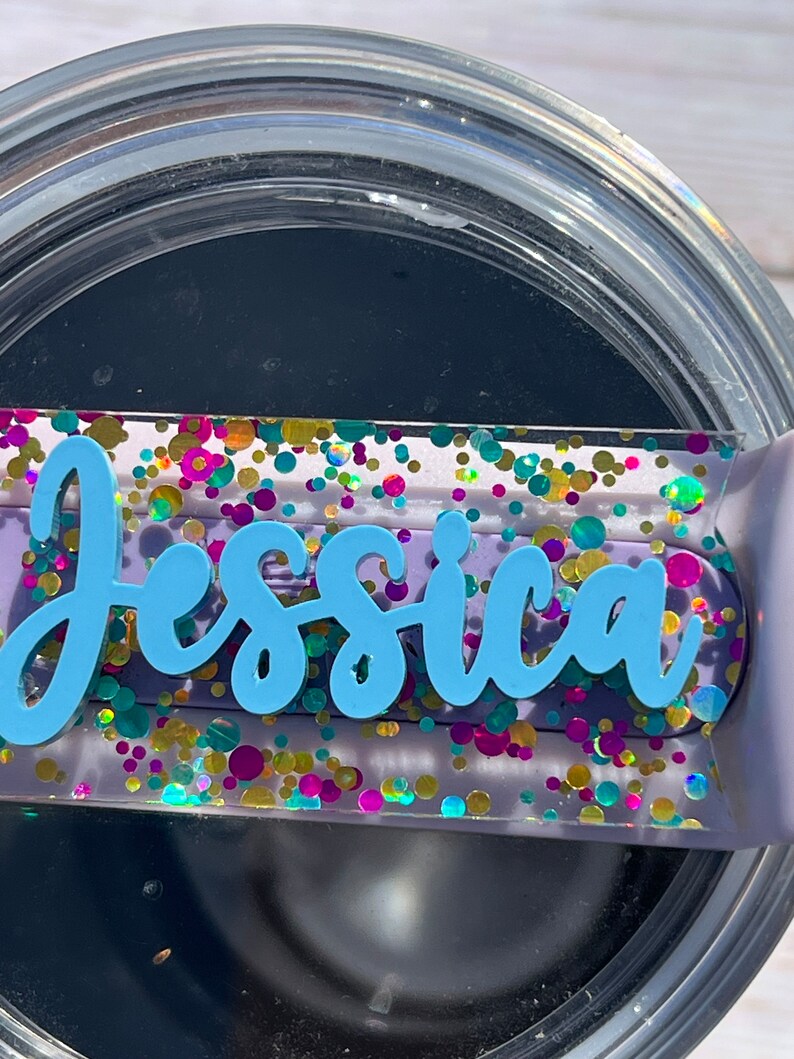 Puede incluir: Un vaso negro con tapa transparente y una etiqueta de nombre personalizada. La etiqueta de nombre est&aacute; decorada con purpurina de colores y el nombre "Jessica" est&aacute; escrito en azul.