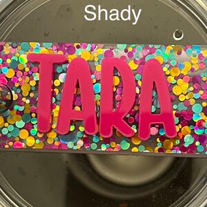 Puede incluir: Un vaso de acero inoxidable con una tapa de purpurina rosa. La tapa tiene un inserto de pl&aacute;stico transparente con el nombre "TARA" en letras rosas. El vaso tiene la palabra "Shady" escrita en el lateral.