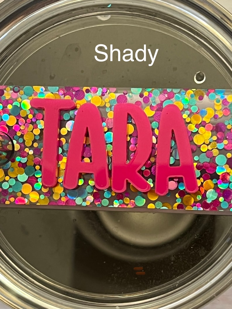 Puede incluir: Un vaso de acero inoxidable con una tapa de purpurina rosa. La tapa tiene un inserto de pl&aacute;stico transparente con el nombre "TARA" en letras rosas. El vaso tiene la palabra "Shady" escrita en el lateral.