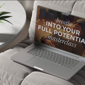 May include: A silver laptop displaying the text "breathe INTO YOUR FULL POTENTIAL masterclass" rests on a grey cushion. A small potted plant with green leaves is on a white table to the left.