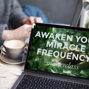 May include: A laptop screen shows the words "AWAKEN YOUR MIRACLE FREQUENCY masterclass" against a green and money background. A cup of coffee on a saucer is next to the laptop. The keyboard is visible.