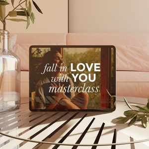 May include: A digital tablet displays the text "fall in LOVE with YOU masterclass" over an image of a person. The tablet rests on a white table with a glass vase holding olive branches. A pink sofa is in the background.