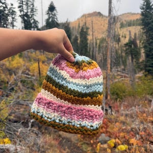 May include: A chunky knit beanie featuring horizontal stripes in shades of pink, cream, light blue, and olive green. The hat is constructed with thick yarn in a ribbed pattern, including a wide folded brim at the base and a gathered crown.