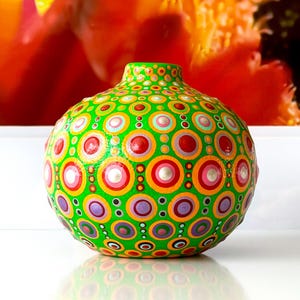 May include: A hand-painted, green ceramic vase with a vibrant pattern of colourful concentric circles. The circles include red, orange, pink, purple, and white accents. The vase has a rounded body and a narrow neck.