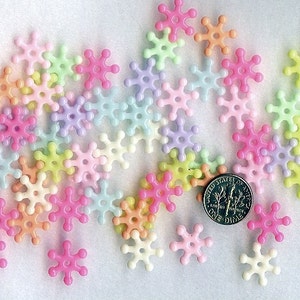 May include: A collection of pastel-colored plastic snowflake-shaped beads. The beads are scattered on a white surface with a US dime for scale.