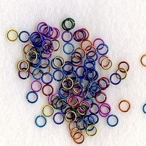 May include: A collection of small, colorful jump rings in various shades of blue, purple, green, gold, and red. These metal rings are scattered on a white surface, suitable for jewelry making or craft projects.