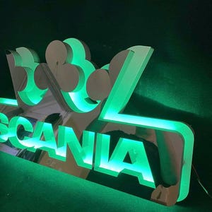 Może przedstawiać: Podświetlany na zielono znak z napisem "SCANIA" i koroną. Znak ma odblaskową powierzchnię i jest podświetlony zielonymi diodami LED. Znak jest prawdopodobnie przeznaczony dla marki ciężarówek lub pojazdów.