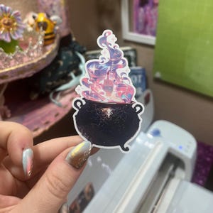 May include: A die-cut sticker featuring a dark purple cauldron with intricate filigree patterns. Rising from the cauldron is a swirling, iridescent cloud of pink, blue, and purple smoke, with a hand emerging from the center showing a middle finger.
