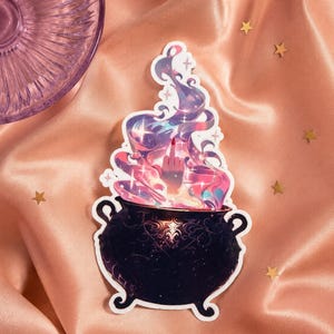 May include: A die-cut sticker featuring a black cauldron with ornate scrollwork patterns. Rising from the cauldron is a swirling cloud of purple, pink, and blue magical smoke, with a hand emerging from the center displaying a middle finger gesture.