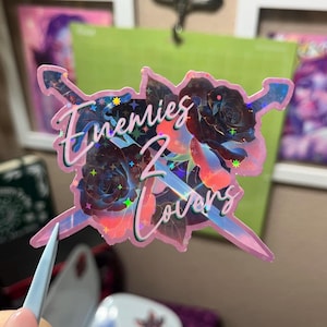 May include: A holographic sticker with the text "Enemies 2 Lovers" in a cursive font, featuring a pink outline and a design of roses and swords. The sticker has a pink border and a shiny, iridescent finish.
