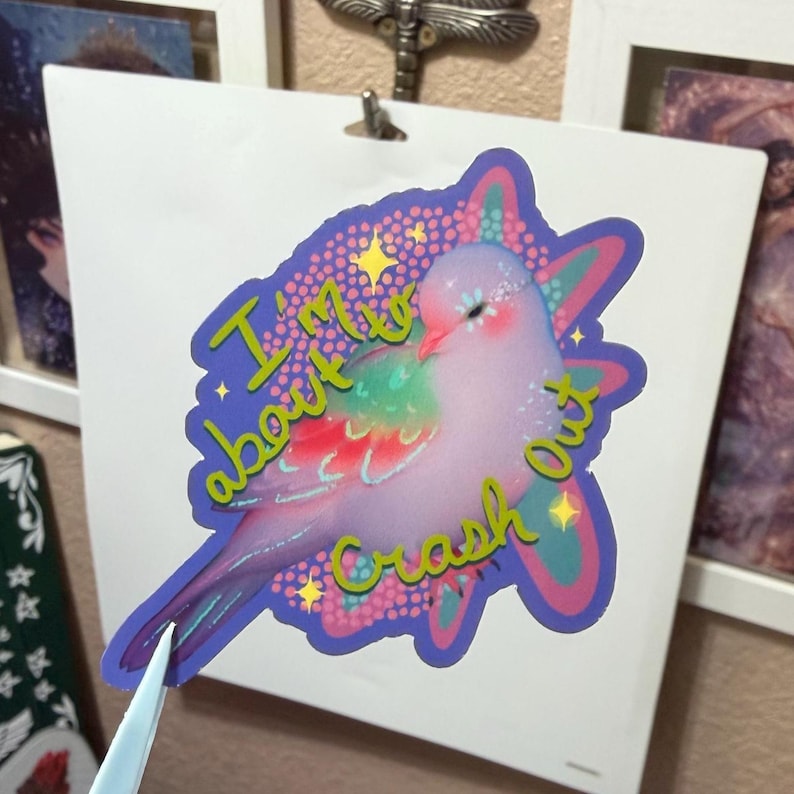 May include: A die-cut sticker featuring a light purple bird with a rainbow-colored wing, surrounded by a pink and teal starburst pattern. The text "I'm about to crash out" is printed in yellow across the bird, accented by small yellow stars.