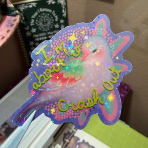 May include: A holographic die-cut sticker featuring a light pink bird with green and red wing accents. The sticker has a purple background with a dotted pattern and yellow text that reads I'm about to crash out, surrounded by small star shapes.