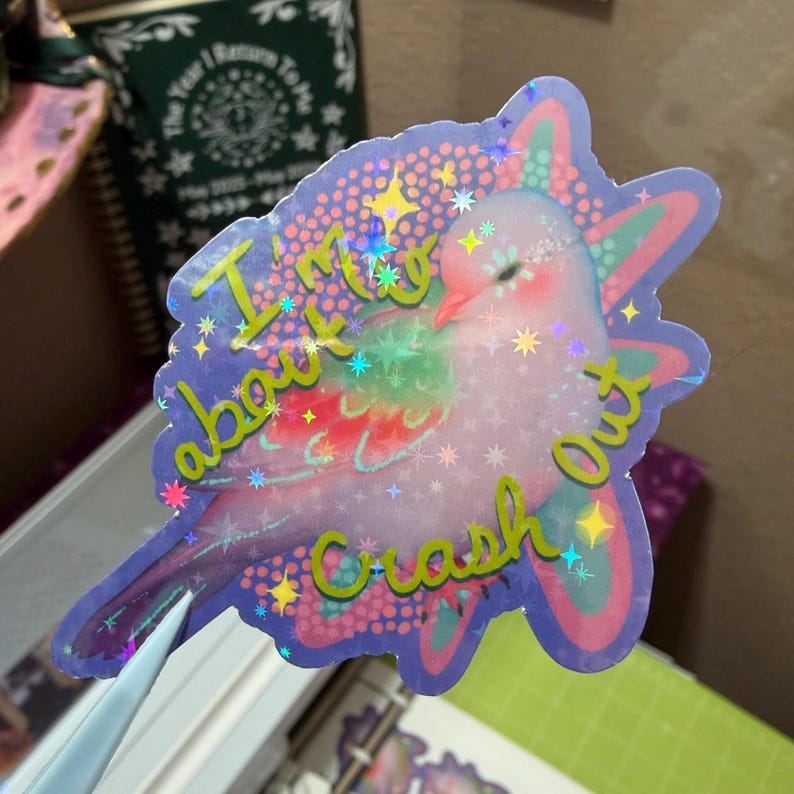 May include: A holographic die-cut sticker featuring a light pink bird with green and red wing accents. The sticker has a purple background with a dotted pattern and yellow text that reads I'm about to crash out, surrounded by small star shapes.