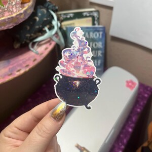 May include: A holographic sticker featuring a black cauldron with ornate detailing and a starry surface. Rising from the cauldron is a swirling, colorful vapor cloud with a hand emerging from the center, displaying a middle finger gesture.