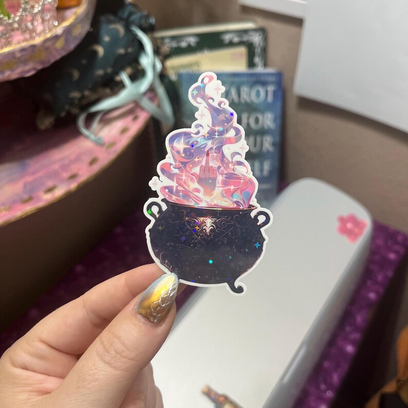May include: A holographic sticker featuring a black cauldron with ornate detailing and a starry surface. Rising from the cauldron is a swirling, colorful vapor cloud with a hand emerging from the center, displaying a middle finger gesture.