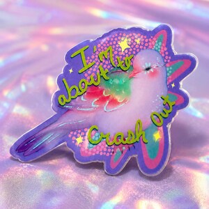 May include: A decorative acrylic pin featuring a light purple bird with iridescent wing accents. The design includes the text 'I'm about to Crash Out' in green lettering, surrounded by small yellow stars and a purple and pink patterned background.
