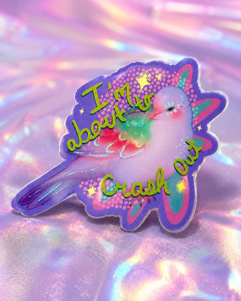 May include: A decorative acrylic pin featuring a light purple bird with iridescent wing accents. The design includes the text 'I'm about to Crash Out' in green lettering, surrounded by small yellow stars and a purple and pink patterned background.