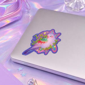 May include: A die-cut sticker featuring a light pink bird with iridescent wings and a tail, surrounded by a purple and blue starburst shape. The text "I'm about to Crash Out" is written in a bright lime green font across the bird and the background.