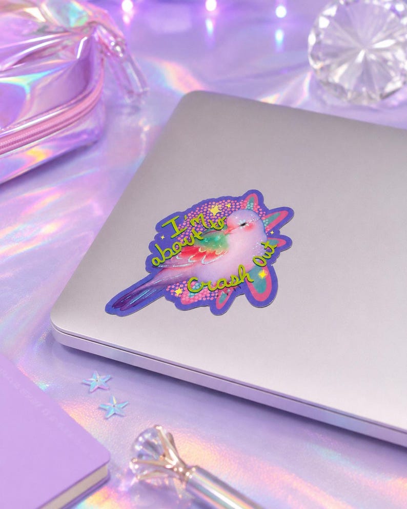May include: A die-cut sticker featuring a light pink bird with iridescent wings and a tail, surrounded by a purple and blue starburst shape. The text "I'm about to Crash Out" is written in a bright lime green font across the bird and the background.