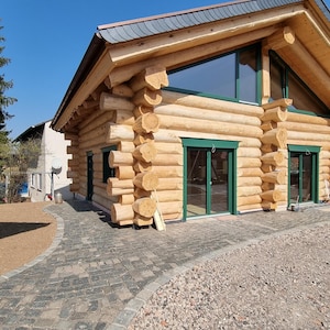 Peut inclure: Un chalet en rondins avec un extérieur brun clair, des encadrements de fenêtres verts et un toit gris foncé. Le chalet a de grandes fenêtres et un ciel bleu clair en arrière-plan. Une allée pavée mène au chalet.