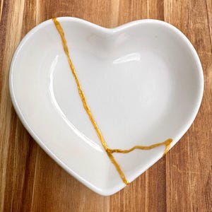 May include: A white, heart-shaped ceramic dish with a gold-colored, decorative line. The dish is on a wooden surface. The gold line appears to be a repair, a nod to the Japanese art of Kintsugi.
