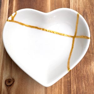May include: A white heart-shaped ceramic dish with gold-colored lines. The dish has a smooth surface and is sitting on a wooden surface. The gold lines are a decorative element.