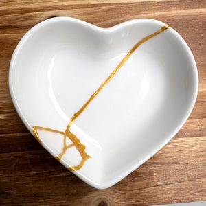 Puede incluir: Un plato de cerámica blanco en forma de corazón con líneas doradas de reparación estilo kintsugi. El plato está sobre una superficie de madera. Las líneas doradas crean un patrón decorativo único en el plato.