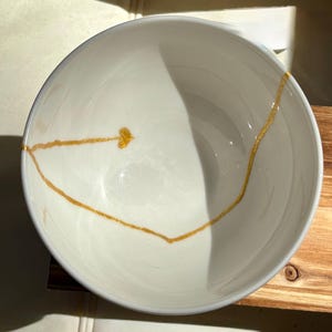 May include: White ceramic bowl with a gold line design. The line is thin and delicate, and it creates a simple and elegant look.