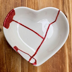 May include: White heart-shaped ceramic dish with red accents, showcasing the Japanese art of Kintsugi. The dish has visible cracks filled with red lacquer, creating a unique and artistic design. The dish is on a wooden surface.