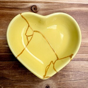 May include: A heart-shaped, yellow ceramic dish with gold-colored kintsugi-style repairs. The dish has a smooth, glossy finish and is set against a wooden background. The gold lines highlight the cracks.