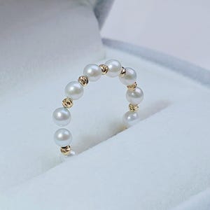 Könnte beinhalten: Ein zarter Ring mit einer Reihe weißer Perlen und goldfarbenen Akzenten. Die Perlen sind gleichmäßig angeordnet und bilden ein geschwungenes Design. Der Ring wird in einer weißen Schmuckschatulle präsentiert, was seine Eleganz unterstreicht.