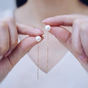 Könnte beinhalten: Ein Paar Ohrhänger. Jeder Ohrring hat einen weißen Perlen-Ohrstecker, der an einer feinen Goldkette befestigt ist. Die Ketten hängen herab und zeigen die Perlen. Der Hintergrund ist weich und unscharf.