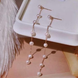 以下が含まれることがあります： 繊細なゴールドチェーンに吊るされた一連の白い真珠が特徴のドロップイヤリング。 イヤリングはポストバック留め具で、白い表面に表示されています。 真珠は丸く、均等に配置されており、エレガントな外観を作り出しています。
