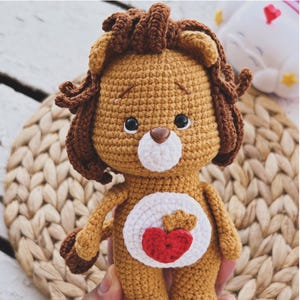 Pode incluir: Um brinquedo amigurumi de leão de crochê feito à mão. O leão é principalmente castanho com uma juba e detalhes castanhos. Possui uma barriga branca com um desenho de coração vermelho. O texto "Care lion" está na parte superior da imagem.