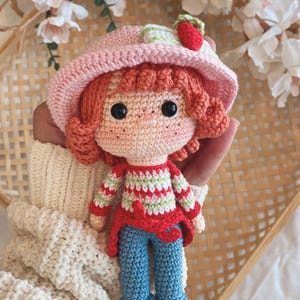 May include: A handmade amigurumi doll, crocheted in the shape of a strawberry girl. The doll wears a pink hat with a strawberry, a striped shirt, a red jacket, and blue pants. The text "Strawberry 2003" is at the top.