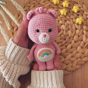 Peut inclure: Un ours rose crocheté avec un arc-en-ciel sur le ventre. L'ours a des yeux noirs, un museau blanc et un nez rose en forme de cœur. L'ours est tenu dans les mains d'une personne, et il y a des bonbons jaunes en forme d'étoile en arrière-plan.