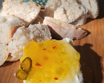 May include: A wooden board with a white cheese topped with orange marmalade and a green pickle. There are pieces of white bread and a blue bowl with green marmalade on the board.