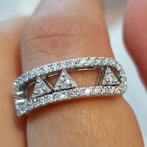 Peut inclure: Gros plan sur une bague en argent au design géométrique. La bague présente trois formes triangulaires au centre, chacune remplie de petites pierres scintillantes. La bande est également incrustée de pierres similaires, créant un effet éblouissant.