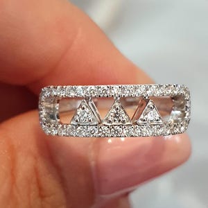 Peut inclure: Une bague en argent avec un design rectangulaire, comprenant trois groupes de diamants triangulaires. La bague est incrustée de petits diamants sur les bords et à l'intérieur des formes triangulaires, créant un effet scintillant. La bague est tenue par une personne.