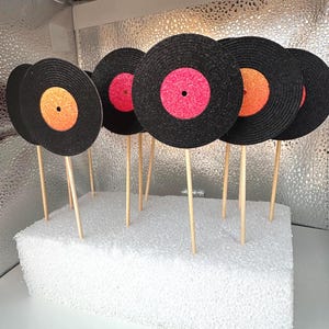 以下が含まれることがあります： オレンジとピンクの中心を持つ、黒い紙製のレコードケーキトッパー8個セット。トッパーは木製の棒に付いており、白い発泡スチロールのベースに配置されています。