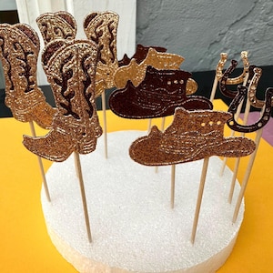 May include: A set of 10 gold glitter cowboy boot, hat, and horseshoe cake toppers. The toppers are on wooden skewers and are ready to be used to decorate a cake.
